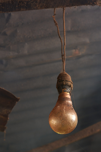 close-up of old dirty or dusty incandescent light bulb and hanging on electric cable with metal roof and smoke in the background, village kitchen lamp in selective focus and copy space