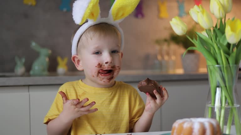 Little cute boy wearing bunny ears eating chocolate easter bunny while sitting