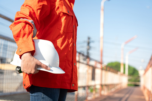 An engineer or construction worker is holding a white safety helmet, wearing orange coverall, standing on the working platform walkway. Ready to work in the challenge workplace concept scene.