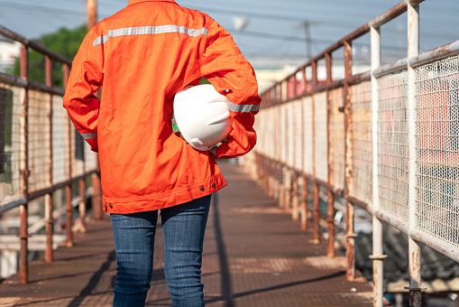 An engineer or construction worker is holding a white safety helmet, wearing orange coverall, standing on the working platform walkway. Ready to work in the challenge workplace concept scene.