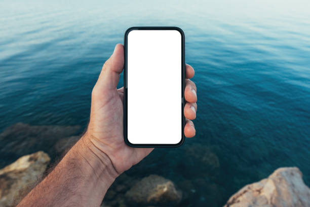 istock Smartphone screen mockup, man holding mobile phone with blank white touchscreen above blue ocean water 1989315501
