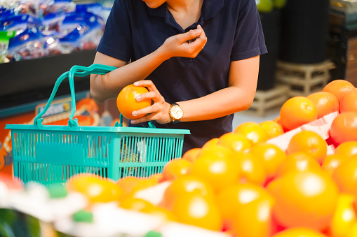 Woman holding grocery basket walk in supermarket. Attractive. female hold orange fruit pick up from shelf for health and wellness in shopping department. Choosing to buy carefully.