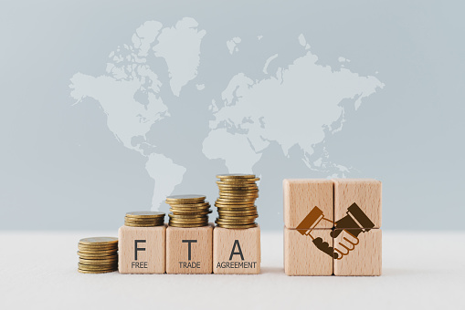 wooden cube blocks with the word FTA, abbreviation for Free Trade Agreement and handshake icon  with stack of coins above on world map background