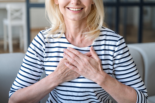 Cropped shot on smiling mature woman holding folded hands on chest, feeling thankful and showing gratitude. Sincere female expressing appreciation with arms on heart. Believe and charity concept