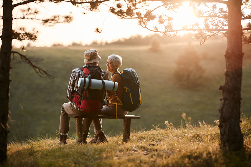 Happy mature couple communicating while taking a break from hiking on a hill at sunset. Copy space.