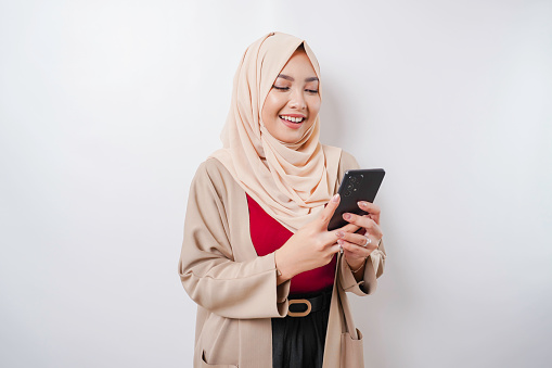 A portrait of a happy Asian woman wearing a hijab and holding her phone, isolated by white background