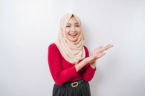 Excited Asian woman wearing hijab pointing at the copy space beside her, isolated by white background