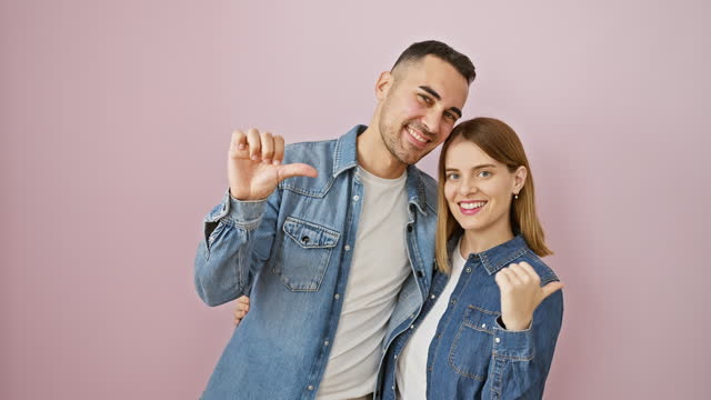 Beautiful couple joyfully pointing behind, thumbs up in confident approval, standing before an isolated pink background, smiling in their chic denim shirts