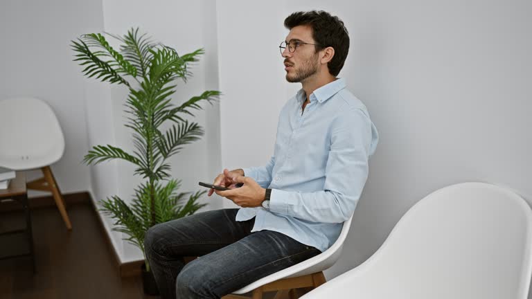 A hispanic man with glasses and beard, using a smartphone while sitting indoors in a white minimalist room.