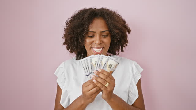 African american woman with curly hair holding us dollars against a pink wall