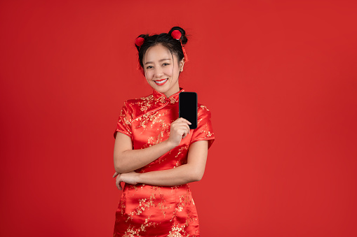 Happy Asian woman wearing qipao dress and showing smartphone isolated on red background.
