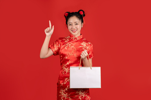 Happy Asian woman wearing red qipao dress holding shopping bag isolated on red background. Happy Chinese new year