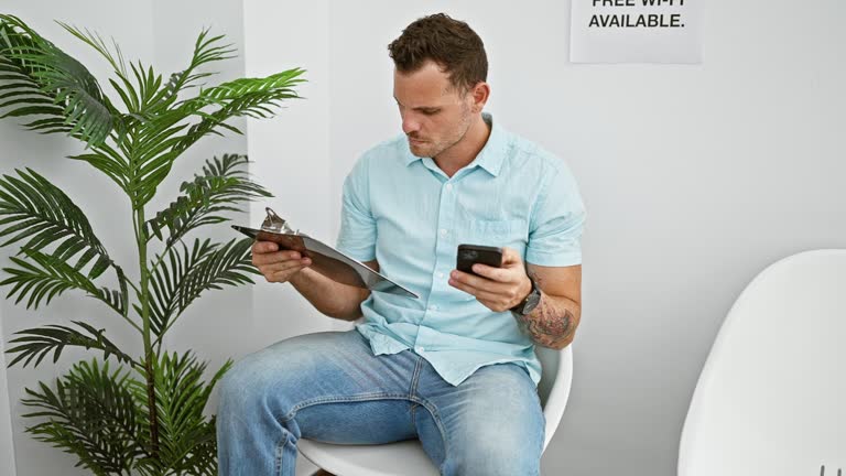 Handsome man with beard using smartphone and holding clipboard in indoor waiting room with plant and chairs.