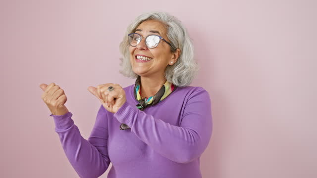 Confident mature woman with grey hair, wearing glasses, giving a thumbs up while pointing behind. smiling wide against an isolated pink background.