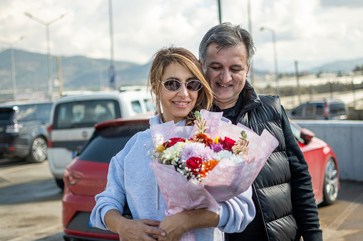 Young woman and her husband standing outdoor by car, holding flowers