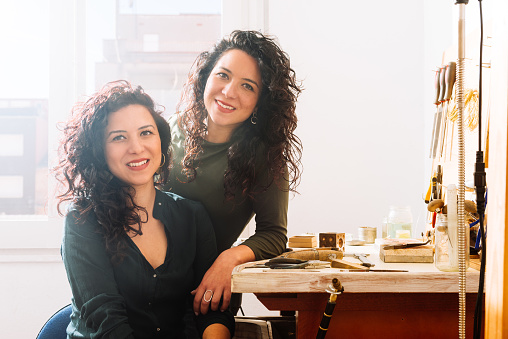 Happy young twin sisters in casual clothes with curly hair smiling and looking at camera while sitting by wooden workbench with tools in daylight at jewelery workshop