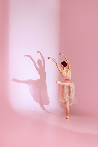 Ethereal Ballet. Classic ballerina dances barefoot in pink traditional ballet dress, tutu against background with her shadow. Concept of elegance, ballet, beauty, action and motion.