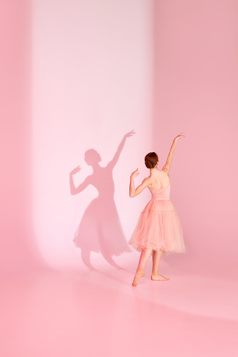 Balletic Grace. Classic ballerina dances barefoot in pink swimsuit and long skirt against pastel pink background with her shadow. Concept of elegance, femininity ballet, beauty, dance school. Ad