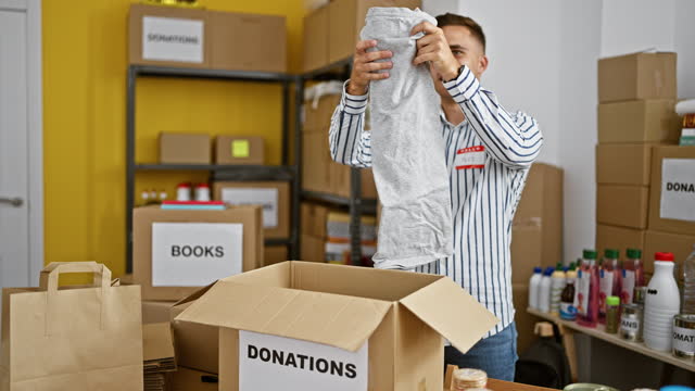 Young hispanic man volunteering in a donation center, organizing clothing and various goods indoors.