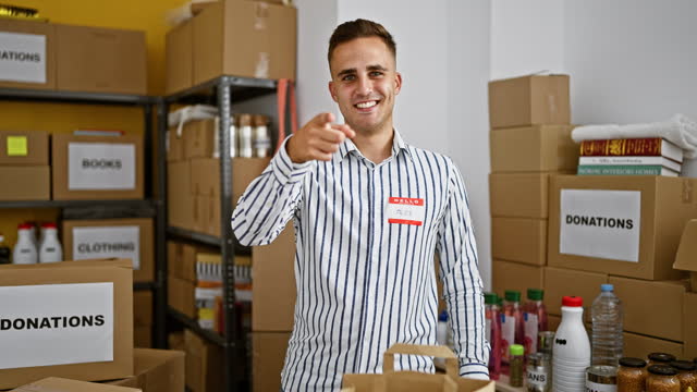 Confident young man with a name tag in a warehouse full of donation boxes looking at the camera.