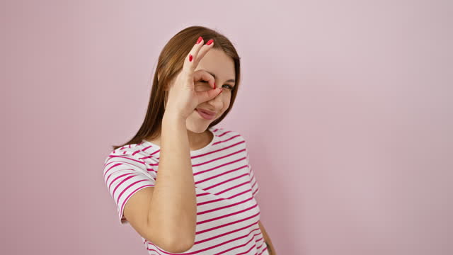 Cheerful young brunette girl in a striped tshirt making fun ok gesture, looking through fingers with a bright smile over pink isolated background