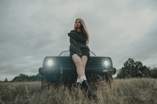 Red-haired girl in green sweater and shorts standing near retro car in nature.