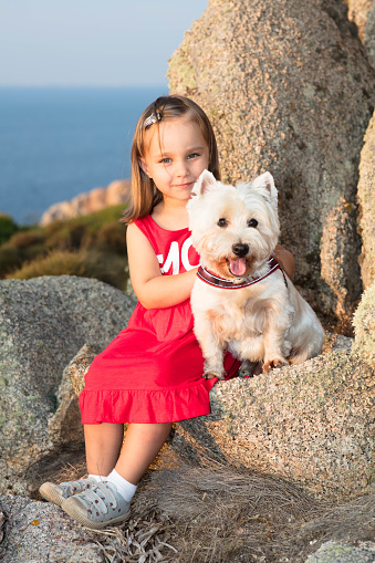 A cute little girl looks happy at sunset, wearing a red dress and holding her little white dog. The guild beam of light from the setting sun gives the portrait a special glow.