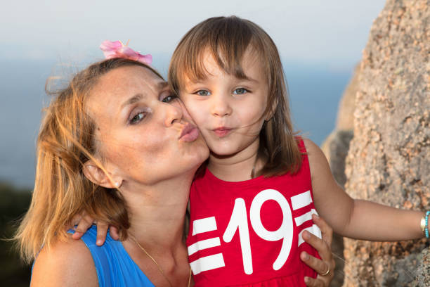 A young beautiful woman looks happy at sunset, wearing a blue shirt and kissing her daughter. The guild beam of light from the setting sun gives the portrait a special glow.