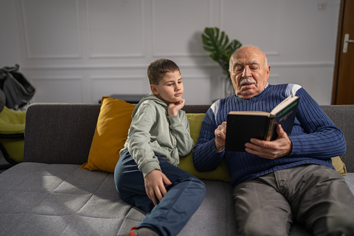 Grandfather and grandson enjoying time together while sitting on sofa and reading a book at home