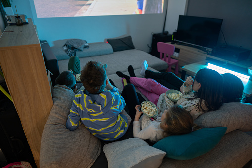 High angle view of group of kids lying on sofa and eating popcorns while watching movie on projector screen at home in the evening