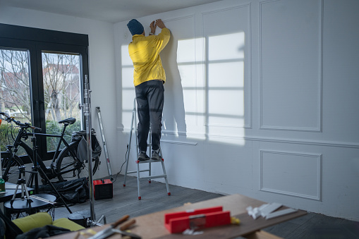 Full length shot of man standing on ladder and setting moldings on white wall during renovation