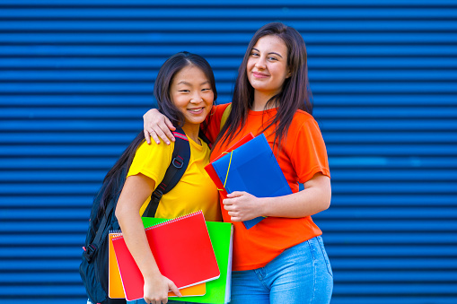 Portrait of two female cute multi-ethnic students embracing in a blue background