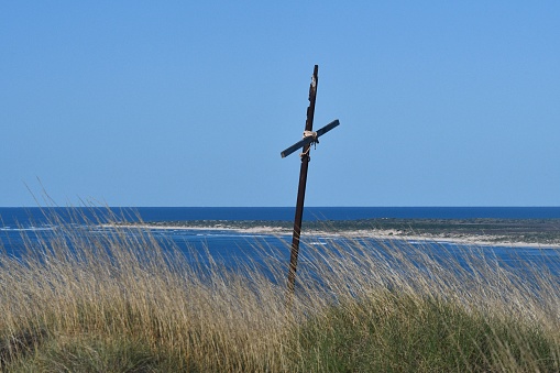 An old wooden cross, tied together with rope, is surrounded by long grass. Behind is a sandy shoreline, and the blue ocean. The sky is clear.