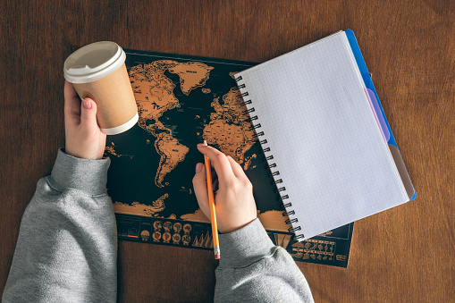 A paper cup in female hands, an empty notepad and a world map on a wooden table. Travel and education concept.
