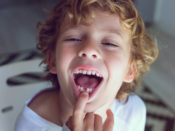 Cute little boy with lost tooth From above of cheerful child with wavy blond hair demonstrating open mouth with lost milk tooth while touching lip and looking at camera baby teeth stock pictures, royalty-free photos & images