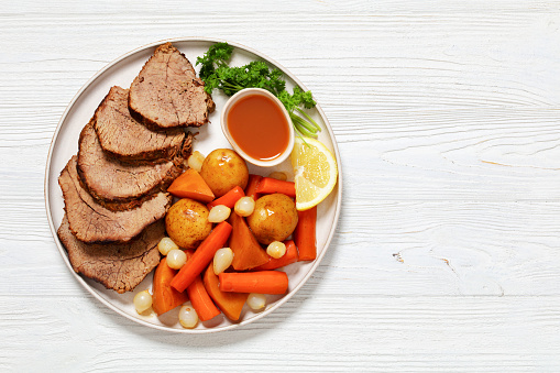 roast beef slices with dipping sauce and winter vegetables, potatoes, carrots, pearl onions, and rutabaga on platter on white wooden table, horizontal view from above, flat lay, free space