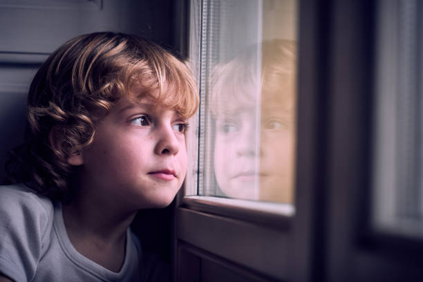 Adorable pensive little boy with curly blond hair leaning on window and looking away dreamily at home