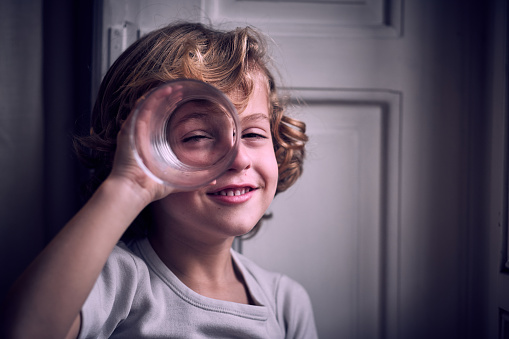 Adorable happy little boy with curly blond hair smiling and looking at camera through glass holding near eye as imaginary binocular