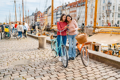 Two female friends with bicycles using a smart phone in Nyhavn Canal in Copenhagen in Denmark.
