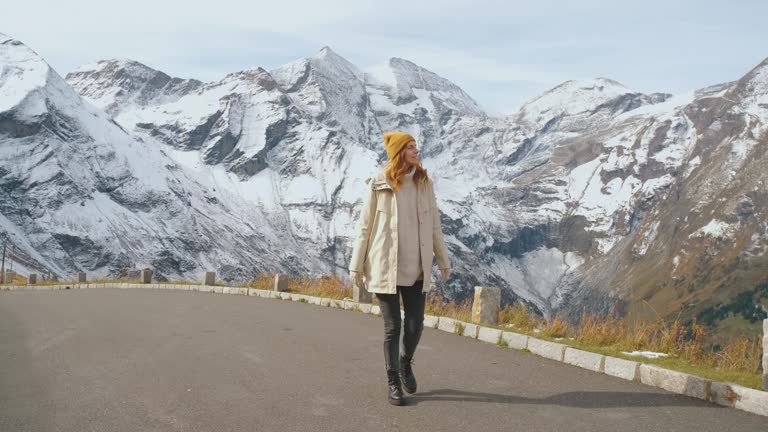 Beautiful girl traveler walks along Majestic Grossglockner Mountain Road in Austria, snow covered sharp peaks of the alpine mountains on background. Incredibly beautiful views in the Alpine mountains. Route in the Alps mountains in autumn or winter. Slow