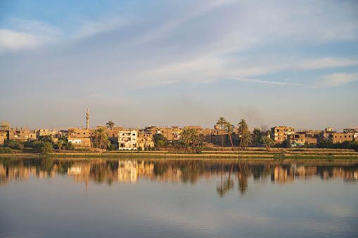 Houses and minaret in warm glow of rising sun reflected in tranquil waters of the River Nile, Luxor, Egypt.