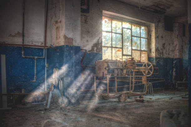 Empty factory hall with peeling walls, exposed beams, and large windows letting in dim light.