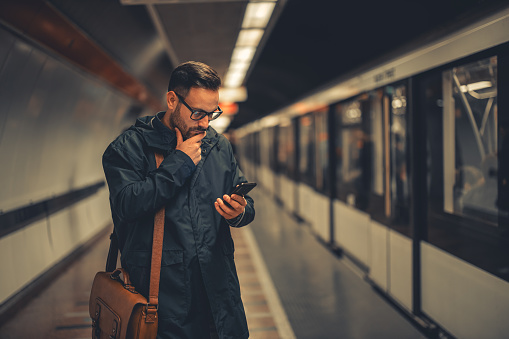 A contemplative adult businessman commuting to work and checking the online train timetable on his smart phone while standing on the subway platform.