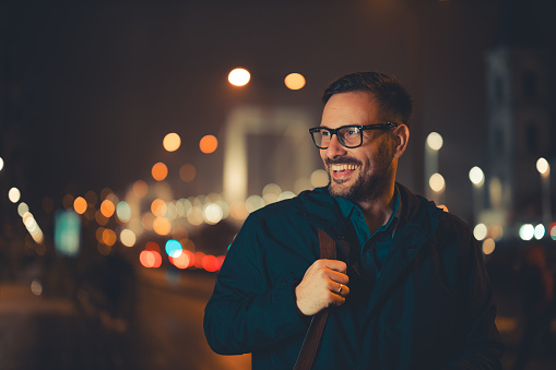 A happy adult businessman with glasses walking back home in the evening and admiring the city around him.