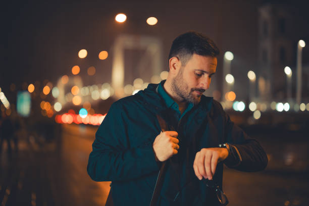 A tired mid adult male employee checking the time on his wristwatch and walking back home during night A displeased mid adult male employee checking the time on his wristwatch after staying late for work and walking back home in the evening. tired after work stock pictures, royalty-free photos & images