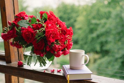 Bouquet of red rose in a vase and a book on a wooden window sill. Still life on the window of an old country house, summer cottage. Floral home decoration.