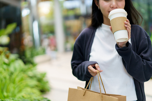 Happy beautiful Asian young woman walking in the shopping mall while she carrying a reusable shopping paper bag and paper takeaway coffee cup on hands. Eco friendly and sustainable lifestyle concept.