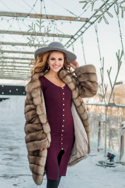 mujer morena que camina por la calle de la ciudad de la nieve con sombrero, vestido rojo y abrigo de piel - russian culture fashion model hat sensuality fotografías e imágenes de stock