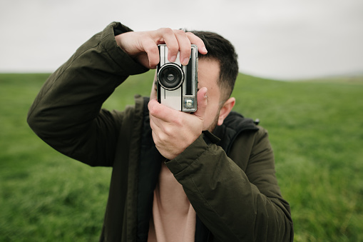 A young man in a green field taking a photo with a vintage film camera
