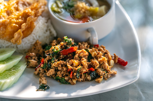 Thai classic spicy menu, Moo Pad Kaprow, or stir-fried ground pork with basil with white rice. Pork basil on a white plate with a Thai omelet over jasmine rice, vegetable soup, and sliced cucumbers.